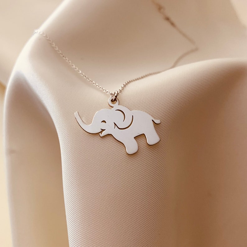 Puede incluir: Un collar de plata con un delicado colgante de elefante. El elefante est&aacute; mirando hacia la derecha y tiene la trompa levantada.