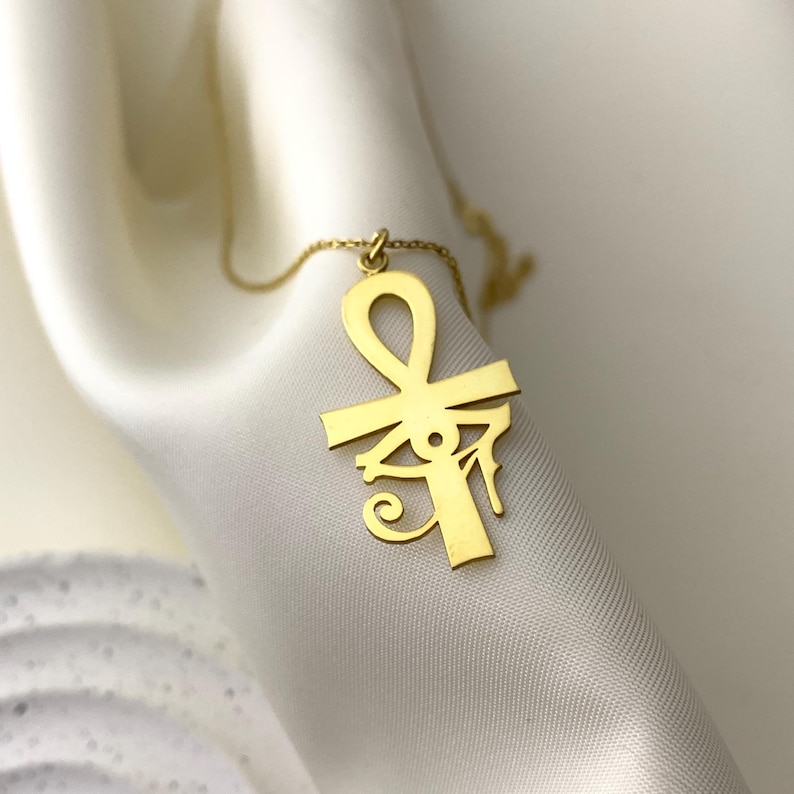 Peut inclure: Un collier pendentif dor&eacute; repr&eacute;sentant un symbole Ankh, une croix avec une boucle en haut, et un design d'&OElig;il d'Horus. Le collier est pr&eacute;sent&eacute; sur un tissu blanc et soyeux.