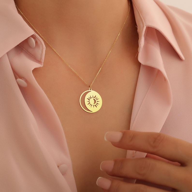 May include: A gold necklace with a pendant featuring a sun and moon design. The pendant is a circle with a crescent moon cut out, and the sun is inside the moon.
