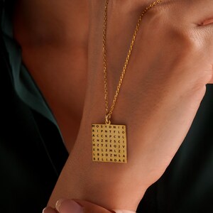 May include: A gold-colored pendant necklace with a square charm. The charm features a word search grid with the name "BELINDAE" visible at the bottom. The necklace is displayed against a dark green background.