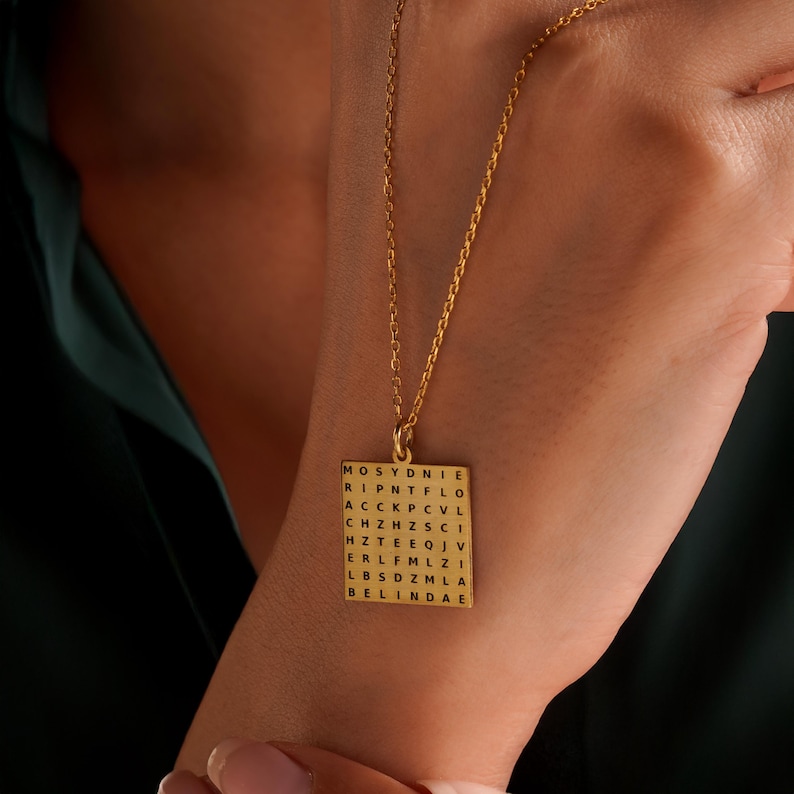 May include: A gold-colored pendant necklace with a square charm. The charm features a word search grid with the name "BELINDAE" visible at the bottom. The necklace is displayed against a dark green background.