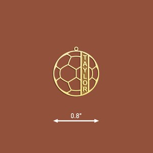 Op de afbeelding: Gouden metalen voetbal ornament met de naam "TAYLOR" verticaal aan de zijkant gegraveerd. Het ornament is 0,8 inch in diameter.