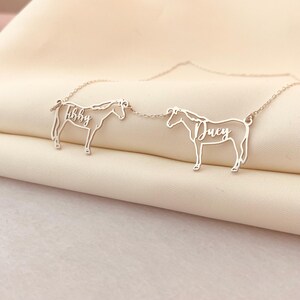 May include: A silver necklace with two horse pendants, each with a name. The horses are linked by a fine chain. One horse has the name "Abby", the other "Duey". The necklace is shown on a light-coloured fabric, with the names in a cursive font.