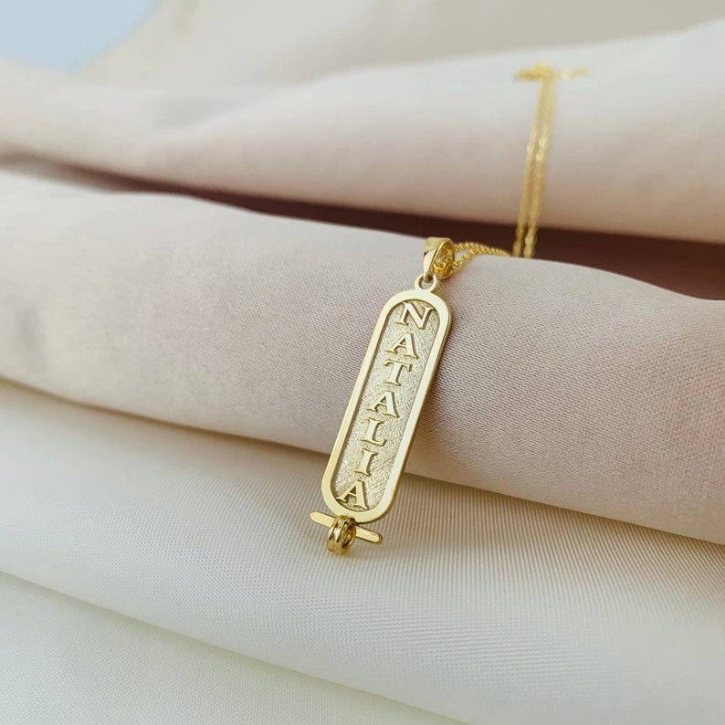 K&ouml;nnte beinhalten: Goldkette mit einem Anh&auml;nger, auf dem "NATALIA" in einer vertikalen, stilisierten Schrift steht. Der Anh&auml;nger ist in Form einer alten &auml;gyptischen Kartusche gestaltet.