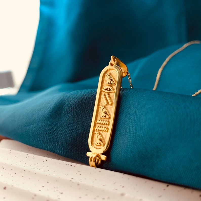 May include: A gold pendant necklace with an Egyptian hieroglyphic design. The pendant features a winged scarab beetle, an eye of Horus, and other symbols.