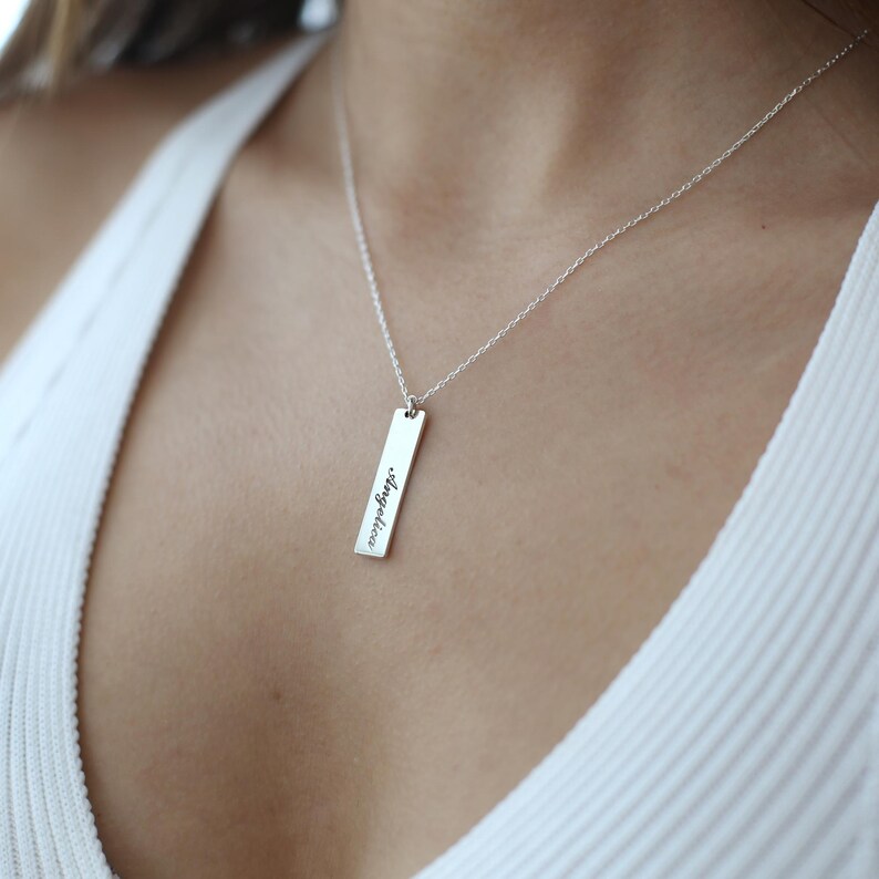 Puede incluir: Un collar de plata con un colgante de barra vertical que dice "Angelica" en cursiva.
