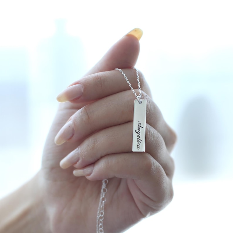 Puede incluir: Un collar de plata con un colgante rectangular que dice "angelica" en escritura cursiva.