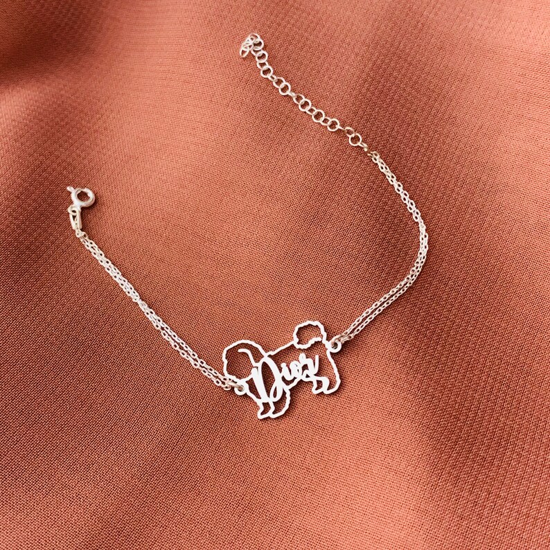 May include: Silver chain bracelet featuring a delicate, cutout design of a dog with the word "Dues" written inside the silhouette.