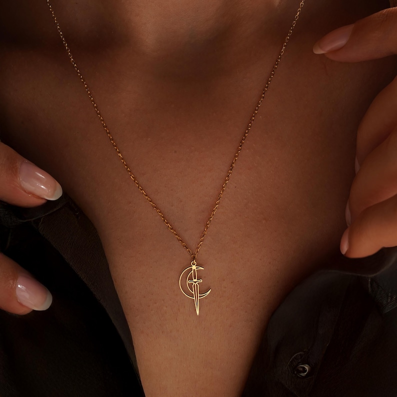 May include: A gold-colored necklace featuring a crescent moon and a sword pendant. The delicate chain and pendant are displayed against a dark background. The necklace is a stylish accessory.