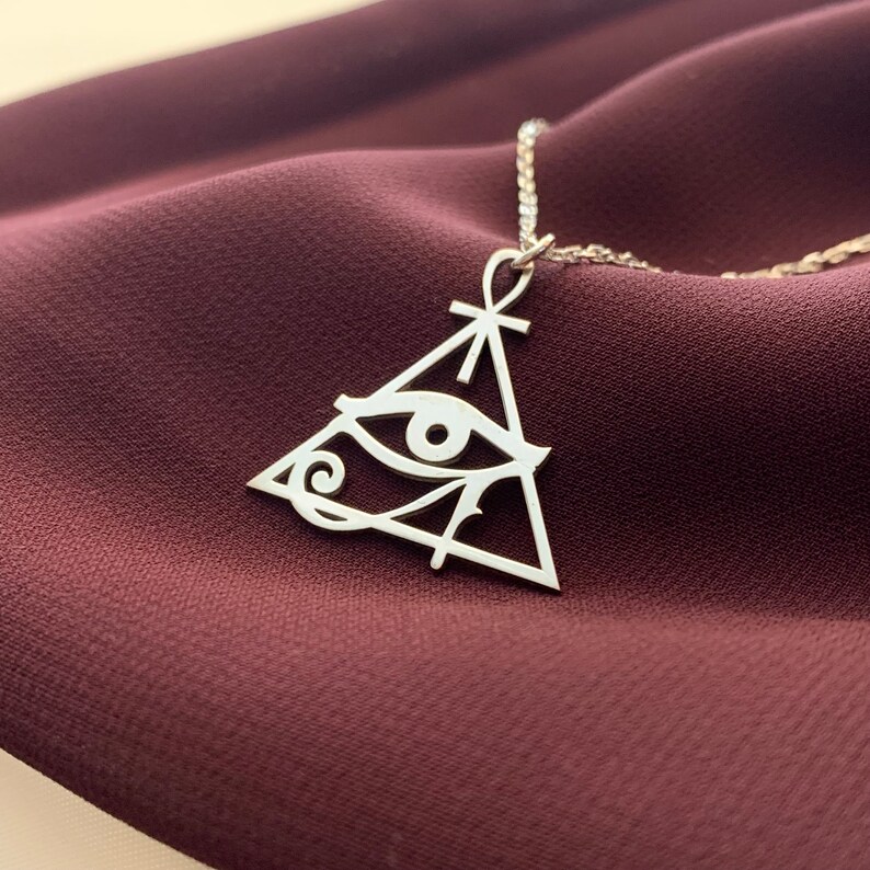 May include: Silver necklace with a pendant featuring an eye of Horus symbol inside a triangle with an ankh on top.