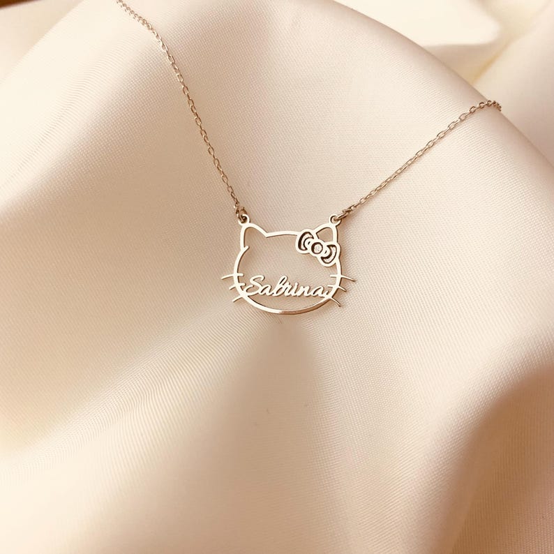 K&ouml;nnte beinhalten: Eine silberne Halskette mit einem Hello-Kitty-inspirierten Anh&auml;nger, in dem der Name "Sabrina" eingraviert ist.