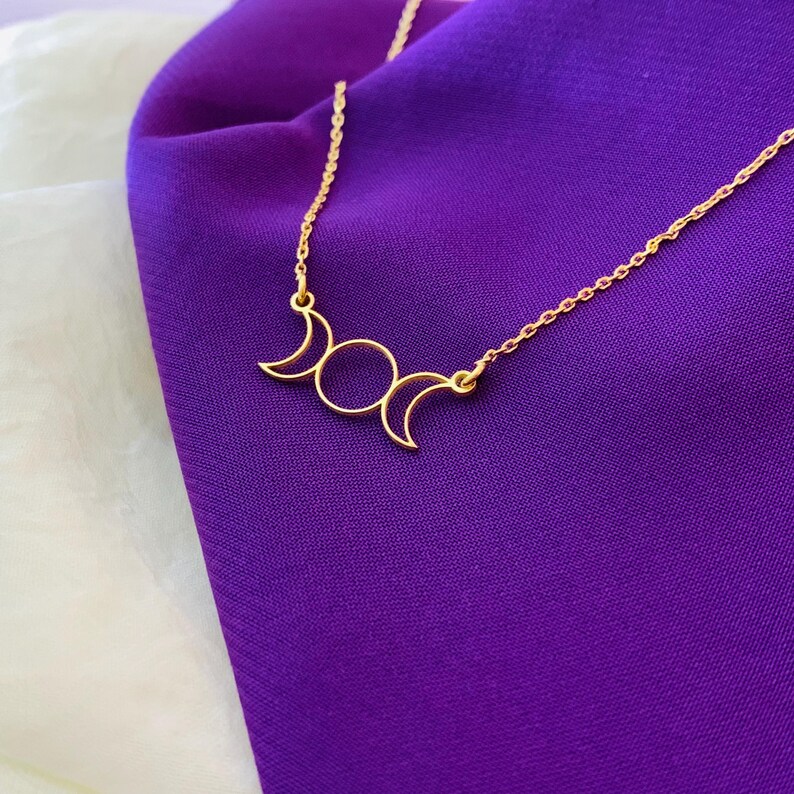K&ouml;nnte beinhalten: Eine goldene Halskette mit einem dreifachen Mond-Anh&auml;nger. Der Anh&auml;nger ist ein schlichtes Design mit drei Mondsicheln.