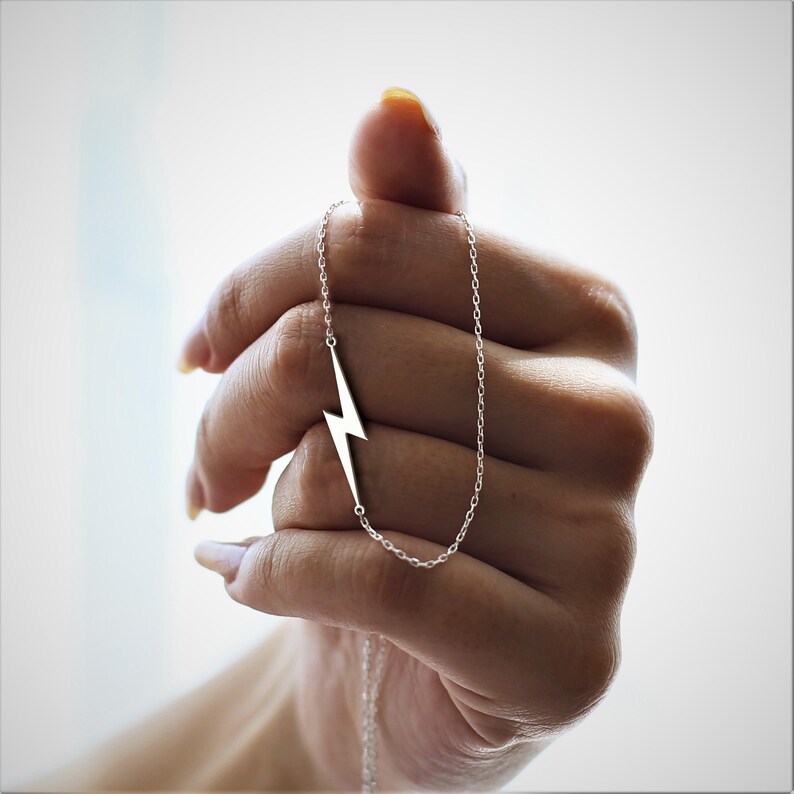 K&ouml;nnte beinhalten: Eine silberne Halskette mit einem Blitz-Anh&auml;nger. Die Kette ist an einer Kette und wird von einer Hand gehalten.
