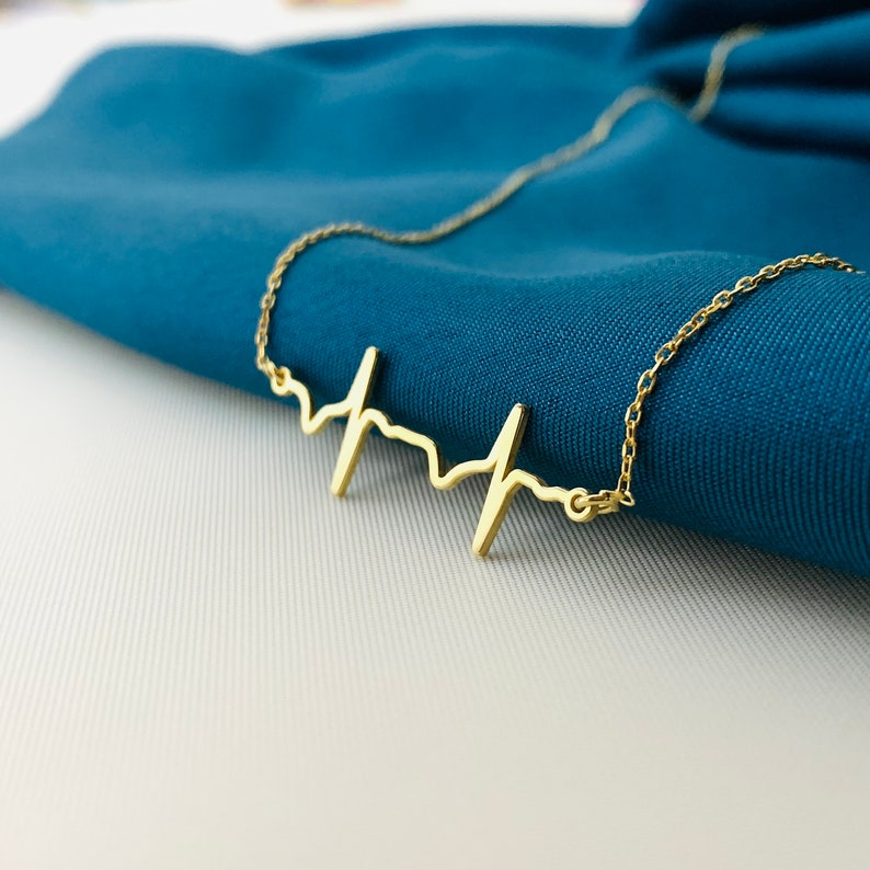 May include: A gold necklace with a heart beat pendant. The necklace is on a blue fabric background.