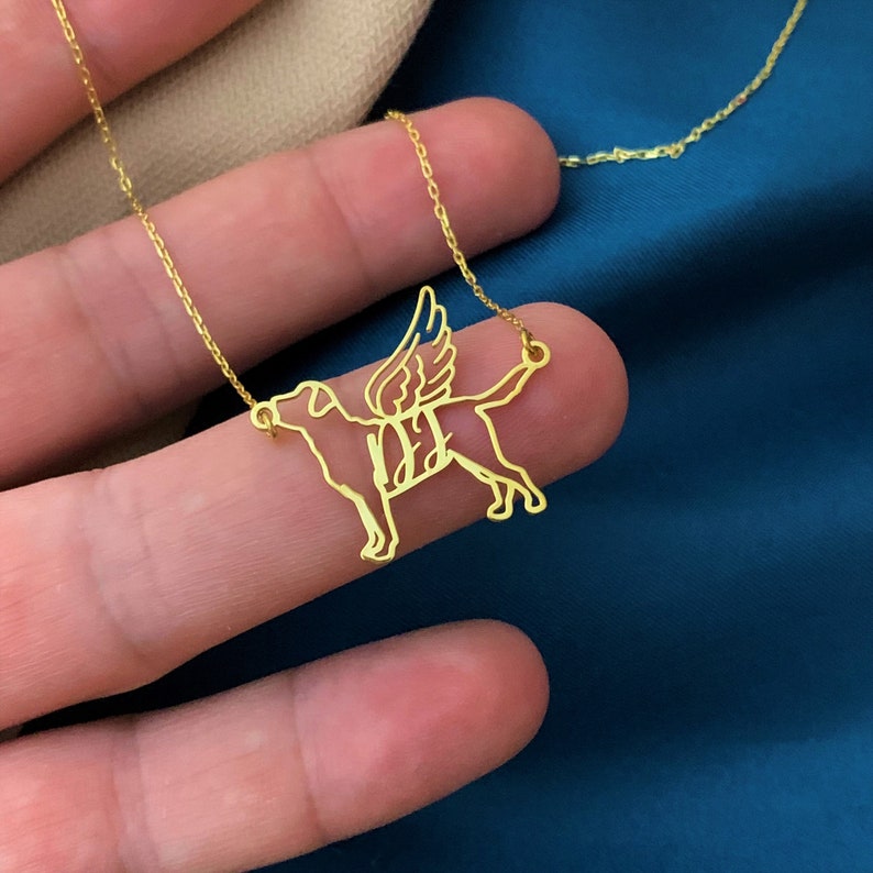 May include: Gold necklace with a pendant of a dog with wings. The dog is outlined in gold and has a delicate design.