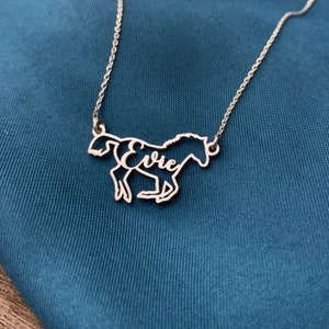 K&ouml;nnte beinhalten: Eine silberne Pferdekette mit dem Namen "Evie", der aus dem K&ouml;rper des Pferdes ausgeschnitten ist. Die Kette liegt auf einem blauen Stoffhintergrund.