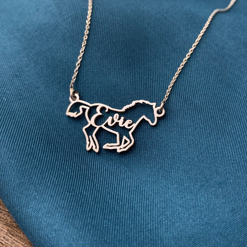 K&ouml;nnte beinhalten: Eine silberne Pferdekette mit dem Namen "Evie", der aus dem K&ouml;rper des Pferdes ausgeschnitten ist. Die Kette liegt auf einem blauen Stoffhintergrund.