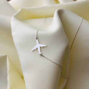 Op de afbeelding: Een zilveren vliegtuig hanger ketting op een witte stoffen achtergrond.