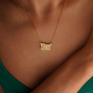 May include: A gold-colored necklace with the phrase "Silence not an Option" in a stylized font. The pendant hangs from a delicate chain. The necklace is displayed on a person wearing a green top.