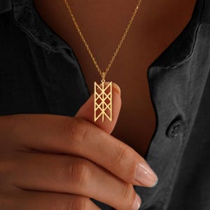 May include: A gold pendant necklace featuring a geometric design. The pendant, suspended from a fine chain, is a rectangular shape with intersecting lines. The necklace is shown against a dark background.