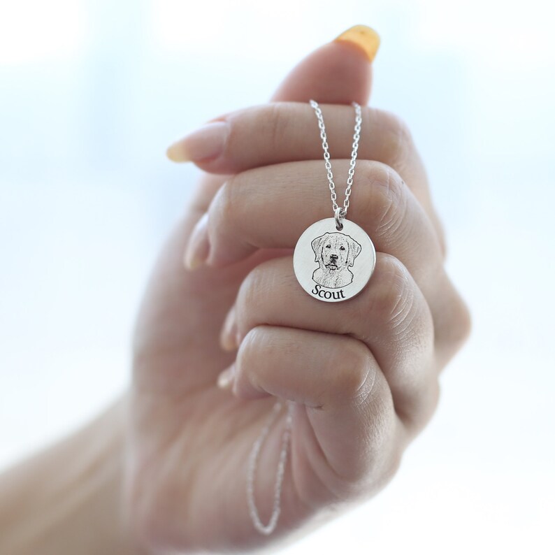 May include: A silver pendant necklace with a detailed engraving of a dog's face. The dog's name, "Scout," is inscribed below the image. The pendant hangs from a delicate silver chain, creating a personalized and sentimental piece of jewellery.