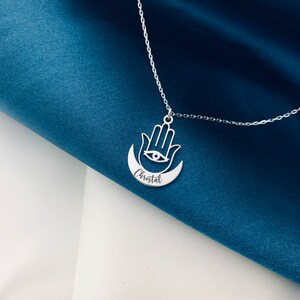 May include: Silver necklace with a hamsa hand charm and a crescent moon pendant. The pendant has an eye in the center and the name "Cristal" engraved on it.