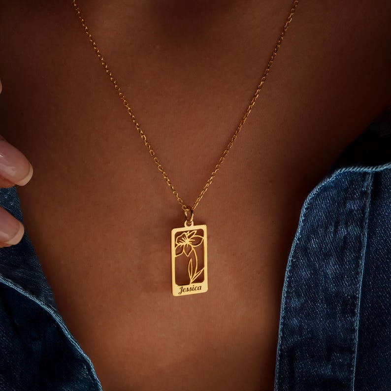 May include: Gold pendant necklace. The rectangular pendant features a floral design and the name "Jessica". The necklace is worn against a denim background.