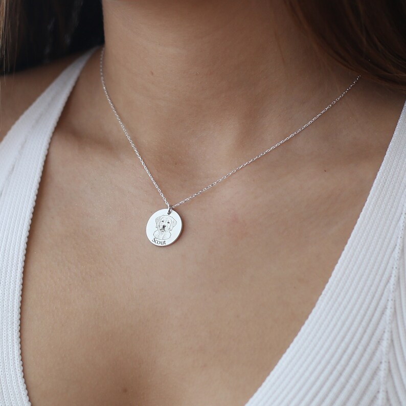 May include: A silver necklace with a circular pendant. The pendant has an engraved illustration of a dog and the name "Scout" below. The pendant hangs from a fine chain, creating a simple and elegant design.