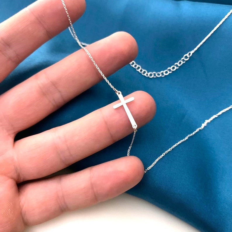 May include: A silver chain necklace with a delicate silver cross pendant. The necklace is on a hand against a blue background.