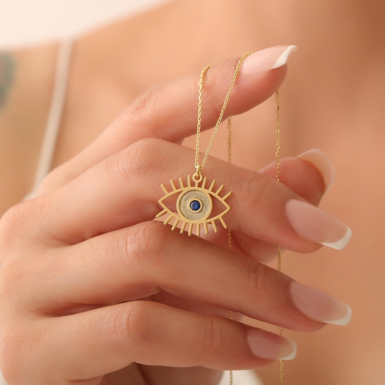 Pode incluir: Um colar de ouro com um pingente de olho azul mau-olhado. O pingente &eacute; um contorno detalhado de um olho com uma pedra preciosa azul no centro.