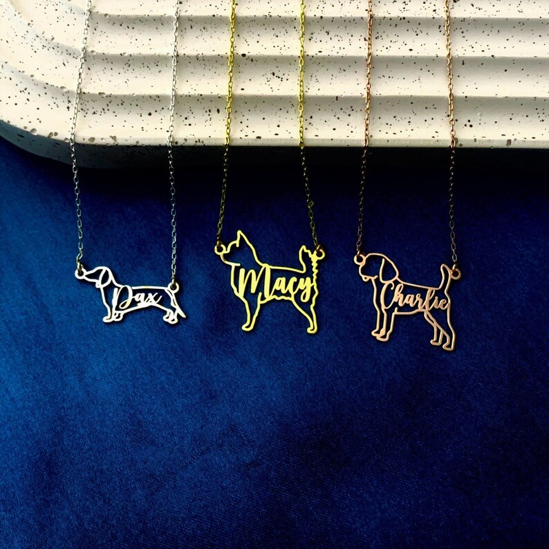May include: Three gold necklaces with dog silhouettes and names. The necklaces are on a white textured surface. The names are "Dax", "Macy", and "Charlie".