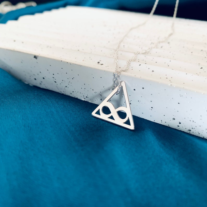 Pode incluir: Um colar prateado com um pingente triangular com um s&iacute;mbolo de infinito vazado. O pingente pende de uma corrente fina. O colar &eacute; exibido em uma superf&iacute;cie branca sobre um fundo azul-petr&oacute;leo.
