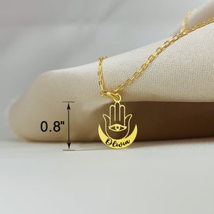 May include: Gold necklace with a hamsa hand charm and the name "Olivia" inscribed on the crescent moon. The charm is 0.8 inches in diameter.
