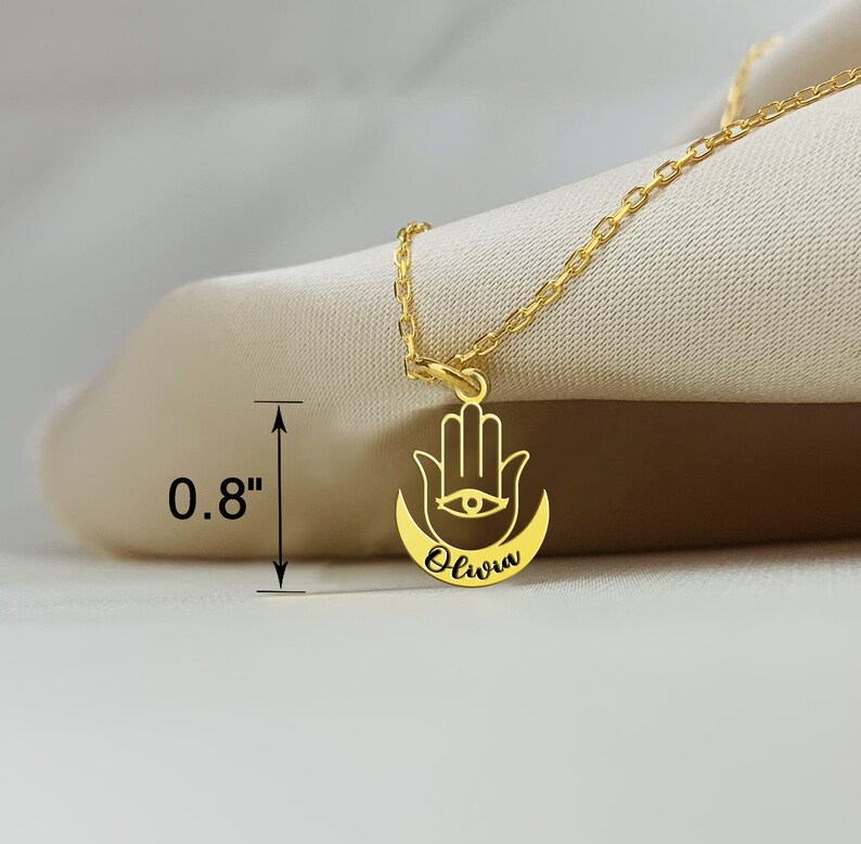 May include: Gold necklace with a hamsa hand charm and the name "Olivia" inscribed on the crescent moon. The charm is 0.8 inches in diameter.
