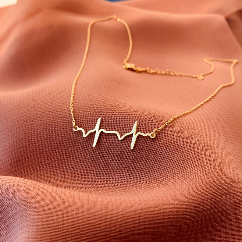 May include: A gold necklace with a delicate heartbeat pendant. The necklace is on a brown fabric background.