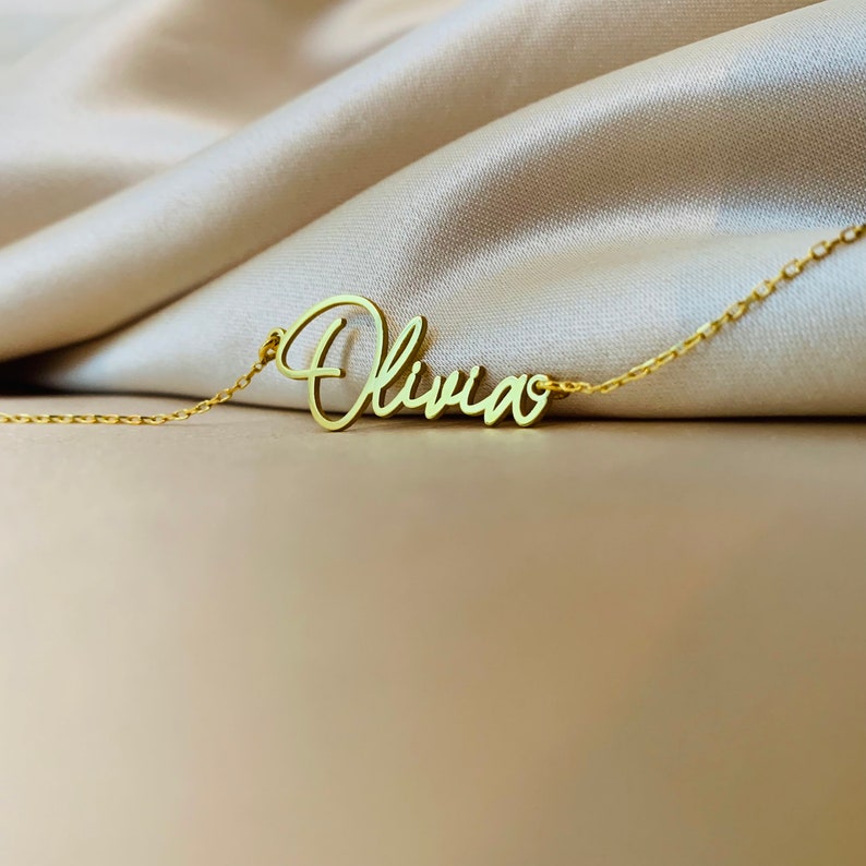 K&ouml;nnte beinhalten: Eine goldene Halskette mit einem Namensschild in Schreibschrift, auf dem "Olivia" steht.