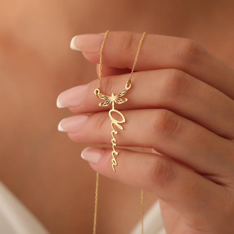 Puede incluir: Collar de oro con un colgante de lib&eacute;lula y el nombre "Olivia" escrito en cursiva.