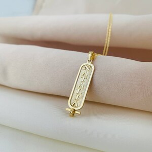 K&ouml;nnte beinhalten: Goldkette mit einem Anh&auml;nger, auf dem der Name "NATALIA" in einer vertikalen, rechteckigen Form eingraviert ist. Der Anh&auml;nger ist so gestaltet, dass er einem alten &auml;gyptischen Hieroglyphen-Cartouche &auml;hnelt.