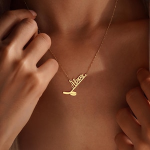 May include: A gold-colored necklace with the name "Alexis" in cursive script, accompanied by a hockey stick and puck design. The necklace is worn around the neck, with the chain visible. The pendant is a delicate piece of jewelry.