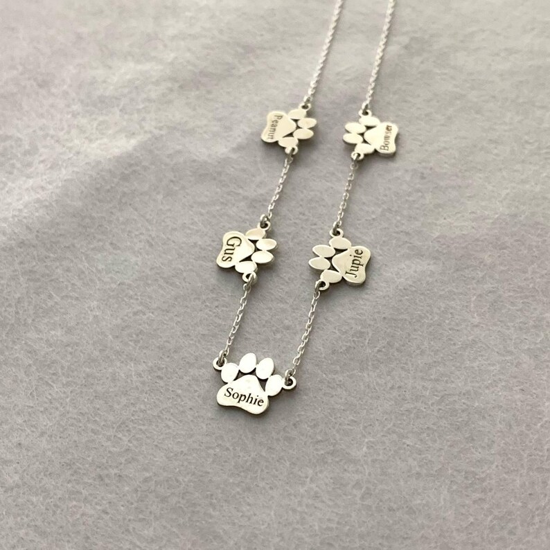 May include: Silver necklace with five paw print charms, each engraved with a different name: Sophie, Gus, Jupie, Bonnie, and Peanut.