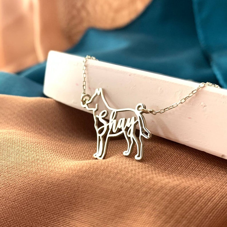 May include: Silver necklace with a dog silhouette pendant. The dog's name, "Shay", is written in cursive inside the silhouette.