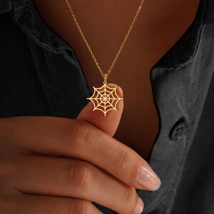 May include: A gold-colored necklace featuring a spiderweb pendant. The pendant is a detailed cutout of a spiderweb design, suspended from a delicate chain. The necklace is shown against a dark background.