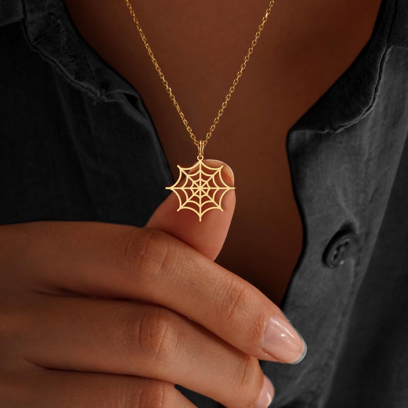 May include: A gold-colored necklace featuring a spiderweb pendant. The pendant is a detailed cutout of a spiderweb design, suspended from a delicate chain. The necklace is shown against a dark background.