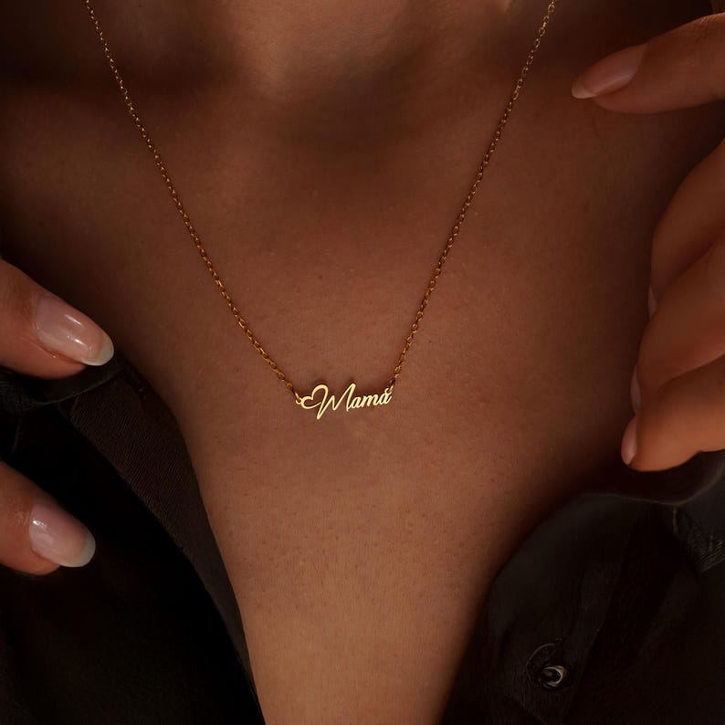 May include: A delicate gold necklace with the word "Mama" in cursive script. The necklace is displayed on a person's neck, with the chain and pendant clearly visible. The pendant is a stylish and sentimental piece of jewelry.