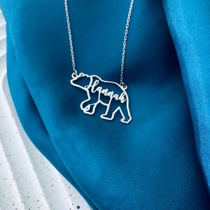 Peut inclure: Collier en argent avec un pendentif en forme d'ours. Le contour de l'ours est d&eacute;coup&eacute; et le nom "Hannah" est &eacute;crit &agrave; l'int&eacute;rieur du corps de l'ours.