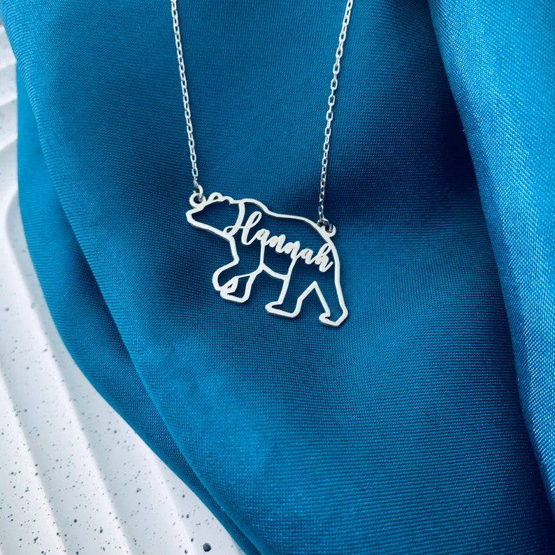 Peut inclure: Collier en argent avec un pendentif en forme d'ours. Le contour de l'ours est d&eacute;coup&eacute; et le nom "Hannah" est &eacute;crit &agrave; l'int&eacute;rieur du corps de l'ours.