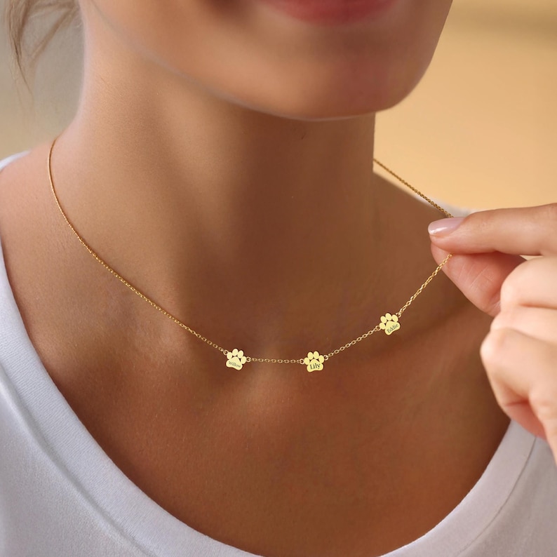 May include: A gold necklace with three paw print charms, each engraved with a name: "Lola", "Lily", and "Sadie".