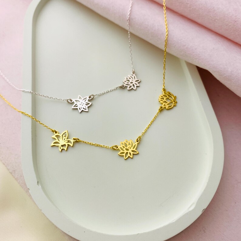 K&ouml;nnte beinhalten: Zwei zarte Halsketten mit Lotusbl&uuml;ten-Anh&auml;ngern. Die obere Halskette ist silberfarben mit drei Anh&auml;ngern, die untere Halskette ist golden mit drei Anh&auml;ngern.