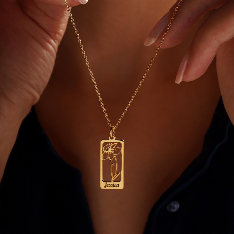 May include: A gold necklace with a rectangular pendant. The pendant features a flower design and the name "Jessica". The necklace is held by a person.