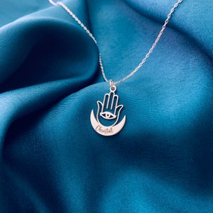 Pode incluir: Colar de prata com um pingente de m&atilde;o de hamsa e um pingente de lua crescente. O pingente tem um olho no centro e o nome "Cristal" gravado na lua.